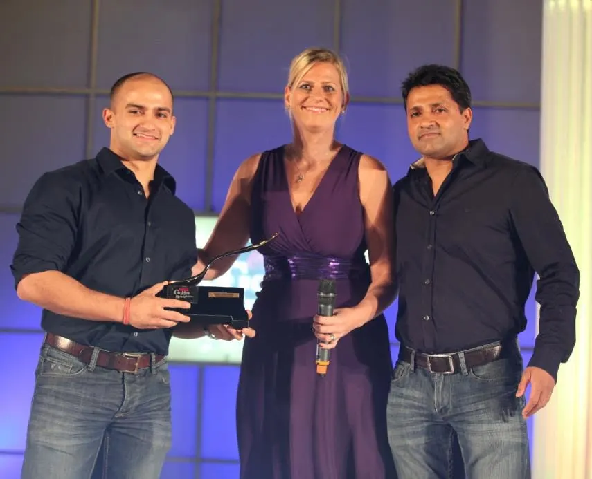Rish Saini, left, on stage holding a black and gold spoon-shaped trophy beside a presenter with a microphone, and his father, Preet Saini, on the right