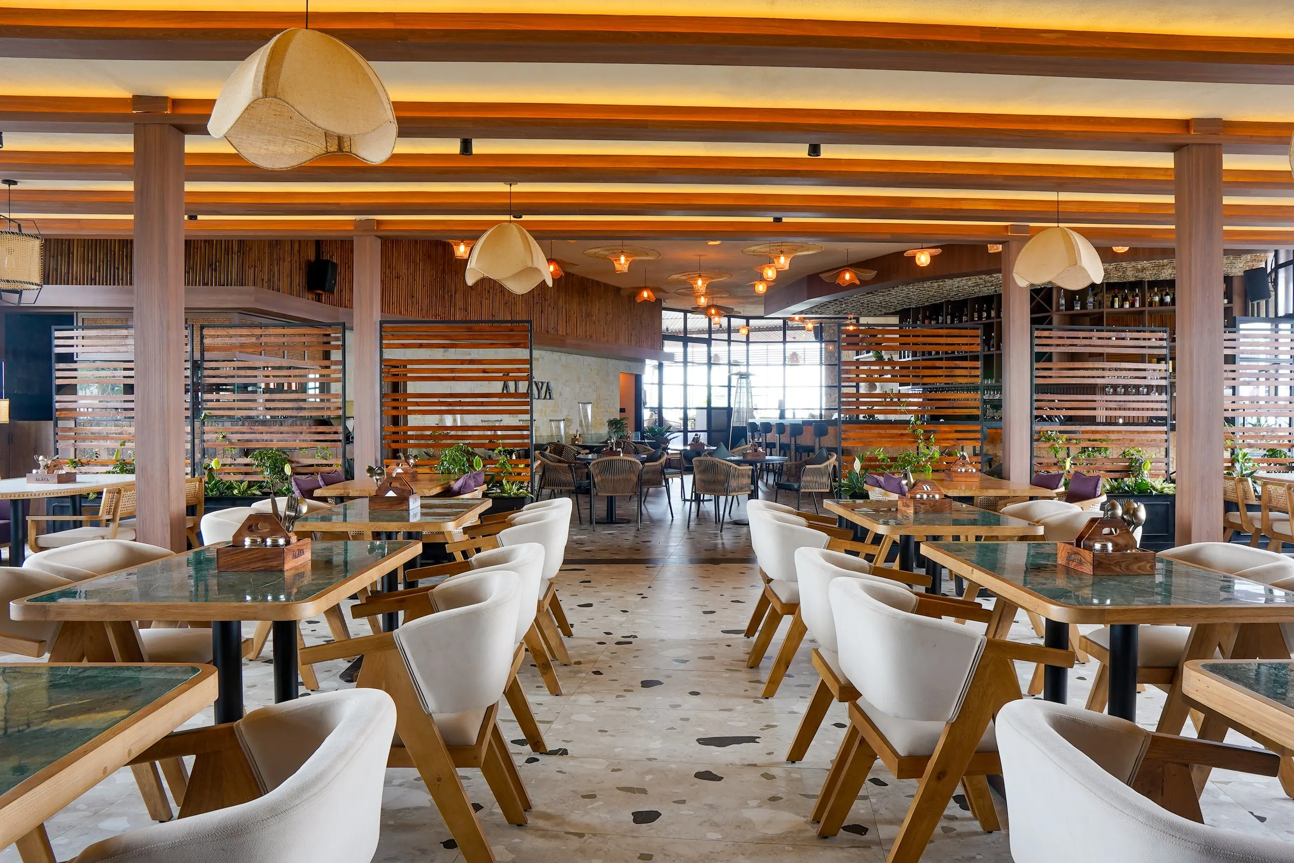 Ālaya's dining hall in Shillong by day: a central aisle between green marble tables and cream chairs, under cove-lit timber beams and fabric pendants, through to the lounge, bar and windows