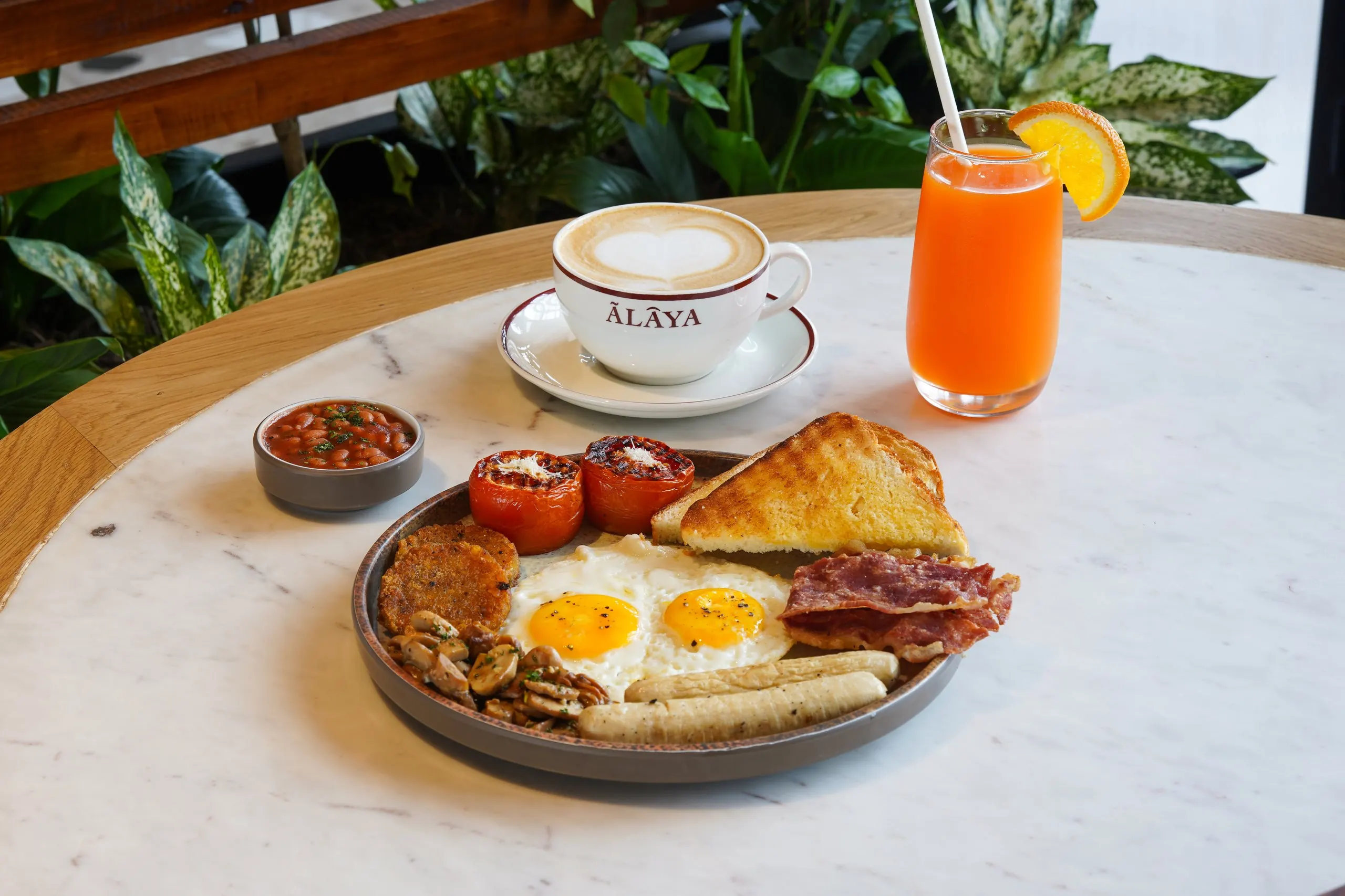 A full breakfast at Ālaya by day: fried eggs, sausages, bacon, toast, grilled tomatoes, hash browns and mushrooms on one plate, with baked beans, a glass of orange juice and a latte in a cup printed with the Ālaya name
