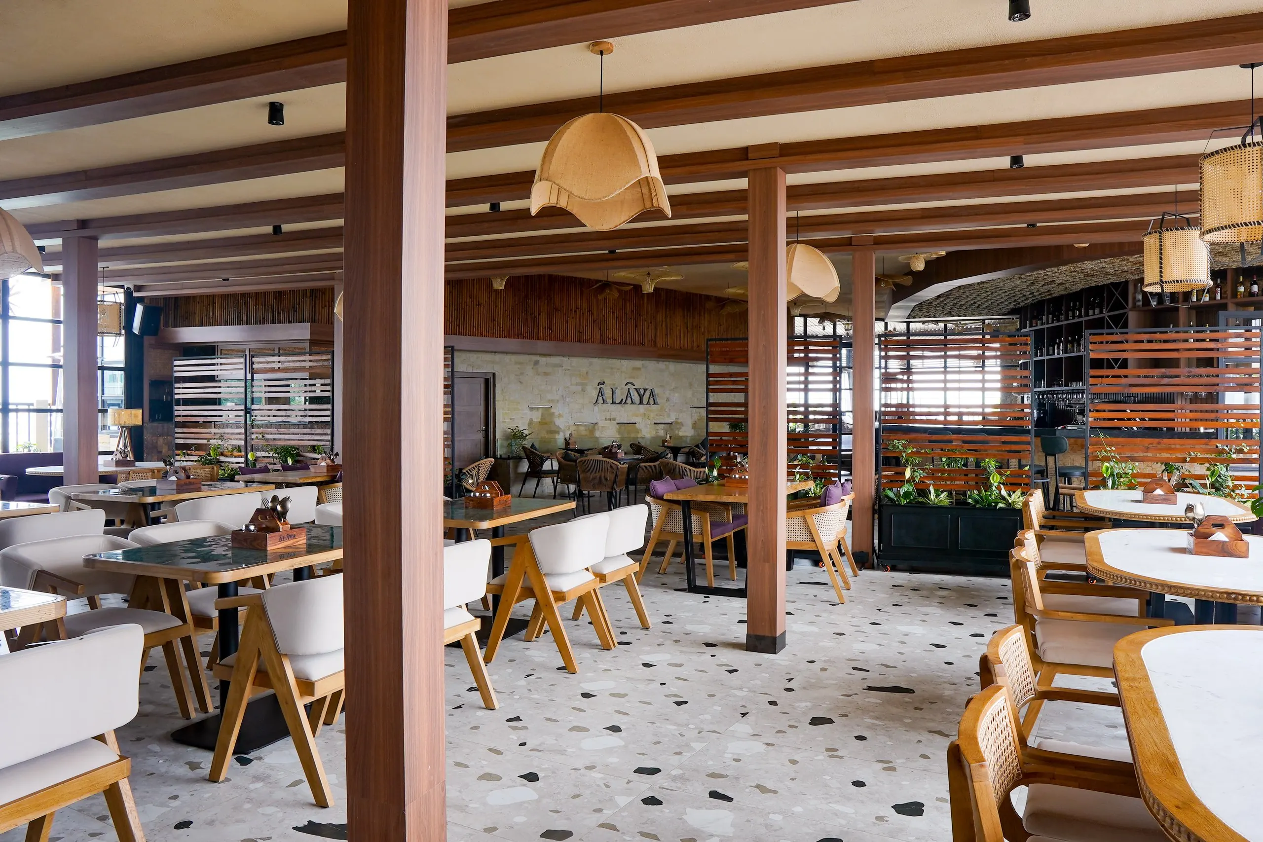 Ālaya's dining room by day, with timber beams, cane furniture and a stone feature wall