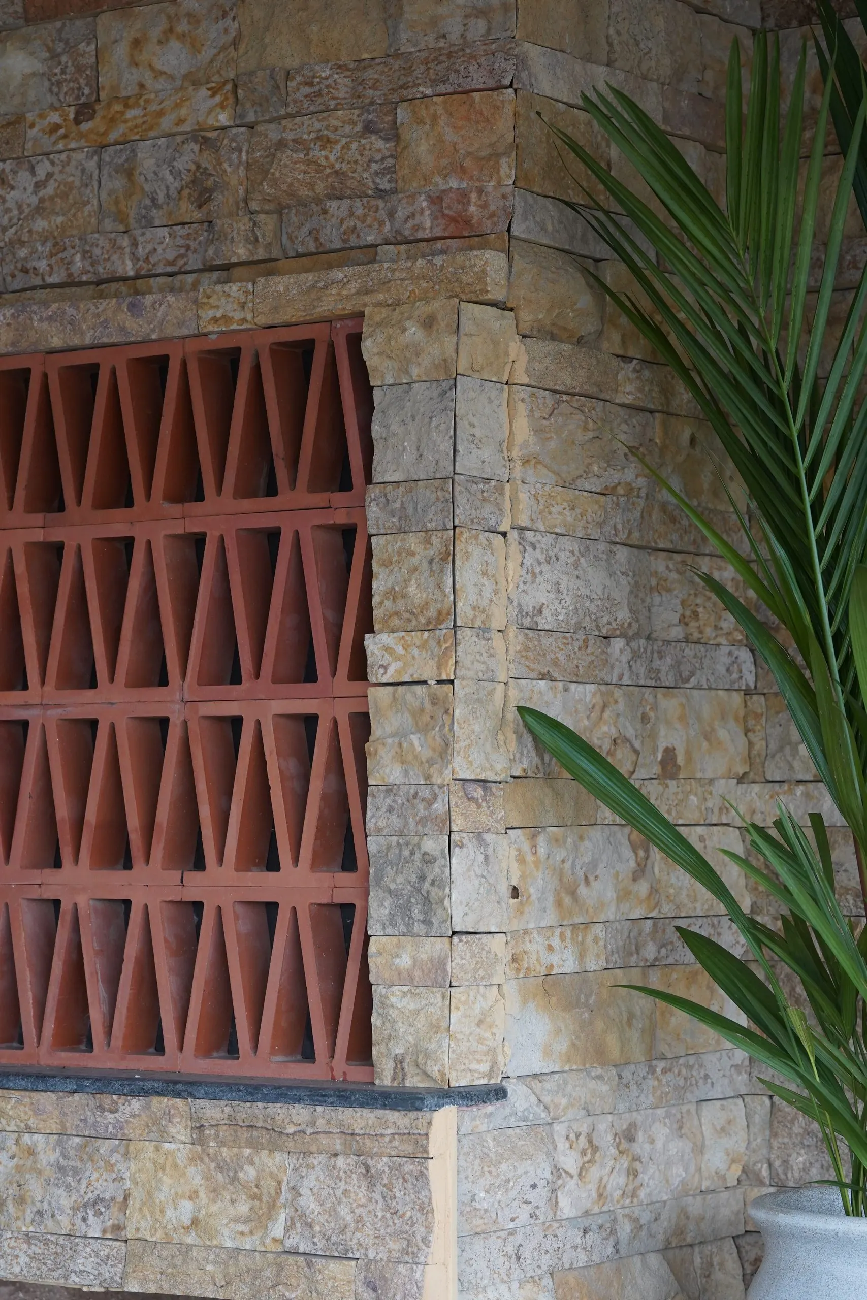 A terracotta jaali screen set into split-face sandstone cladding at Ālaya, beside a palm