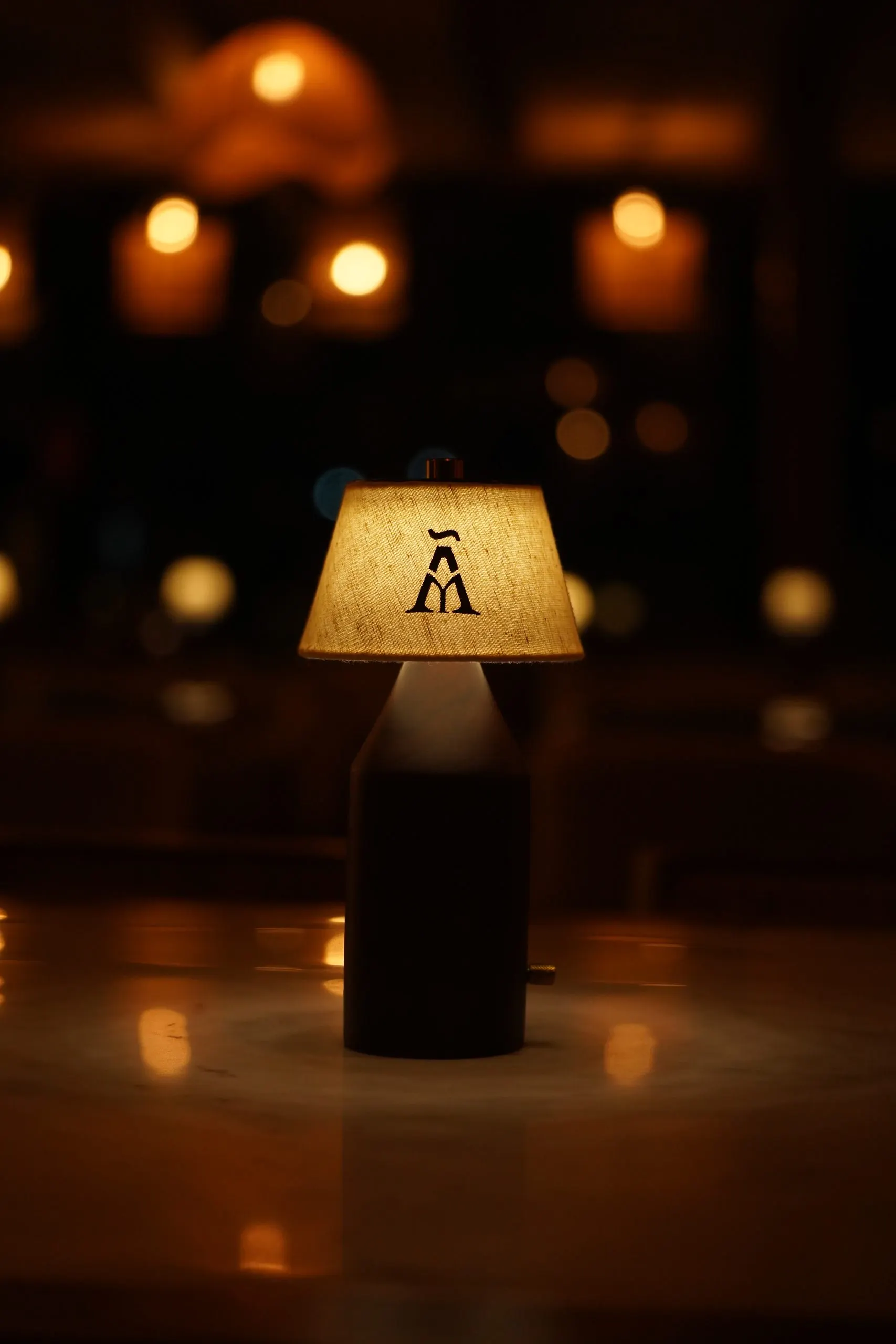 A table lamp with the Ālaya monogram on its shade, lit on a table at night