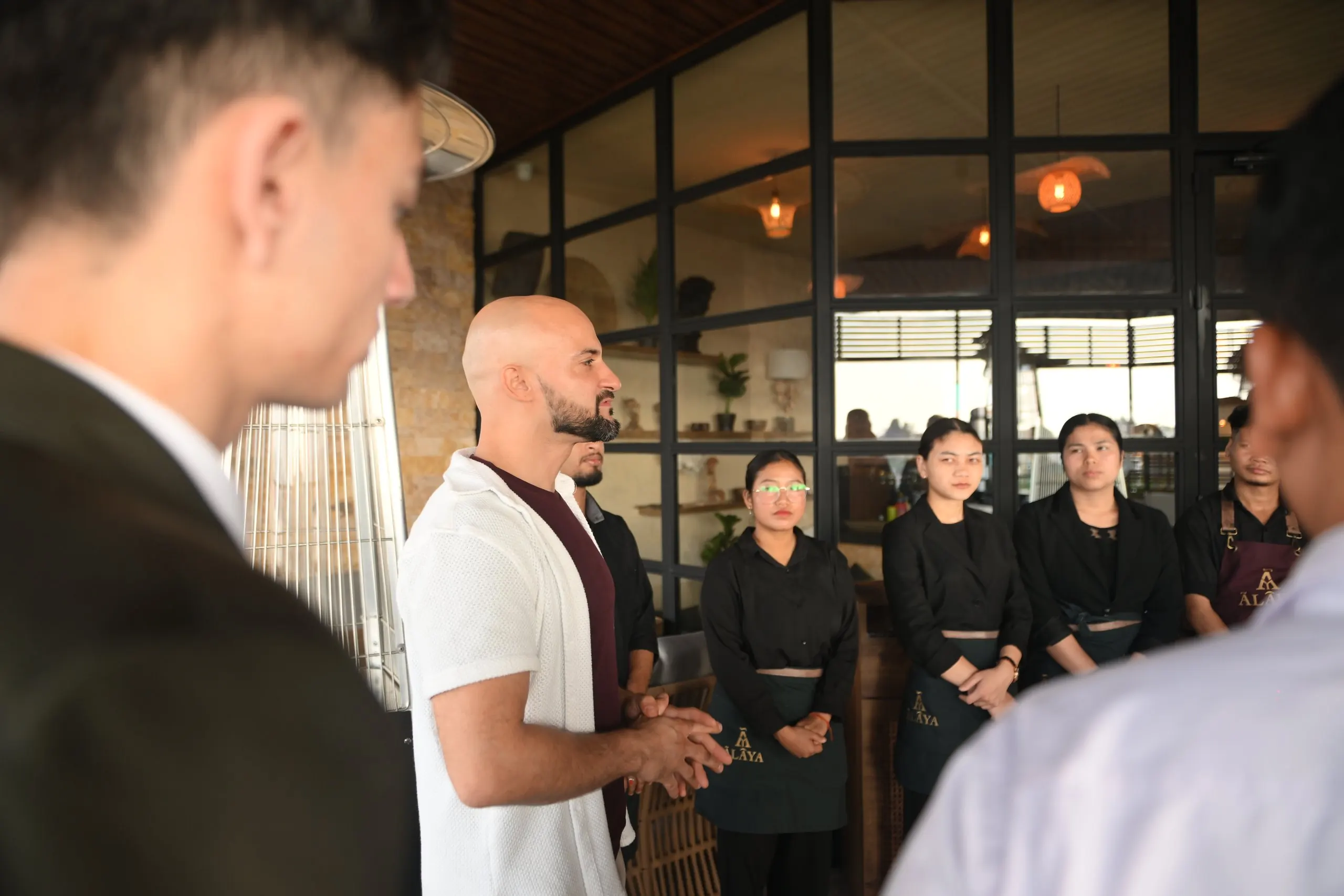 A briefing for the floor team on the Ālaya terrace by day: a man in a white overshirt speaks to staff in black uniforms and Ālaya aprons