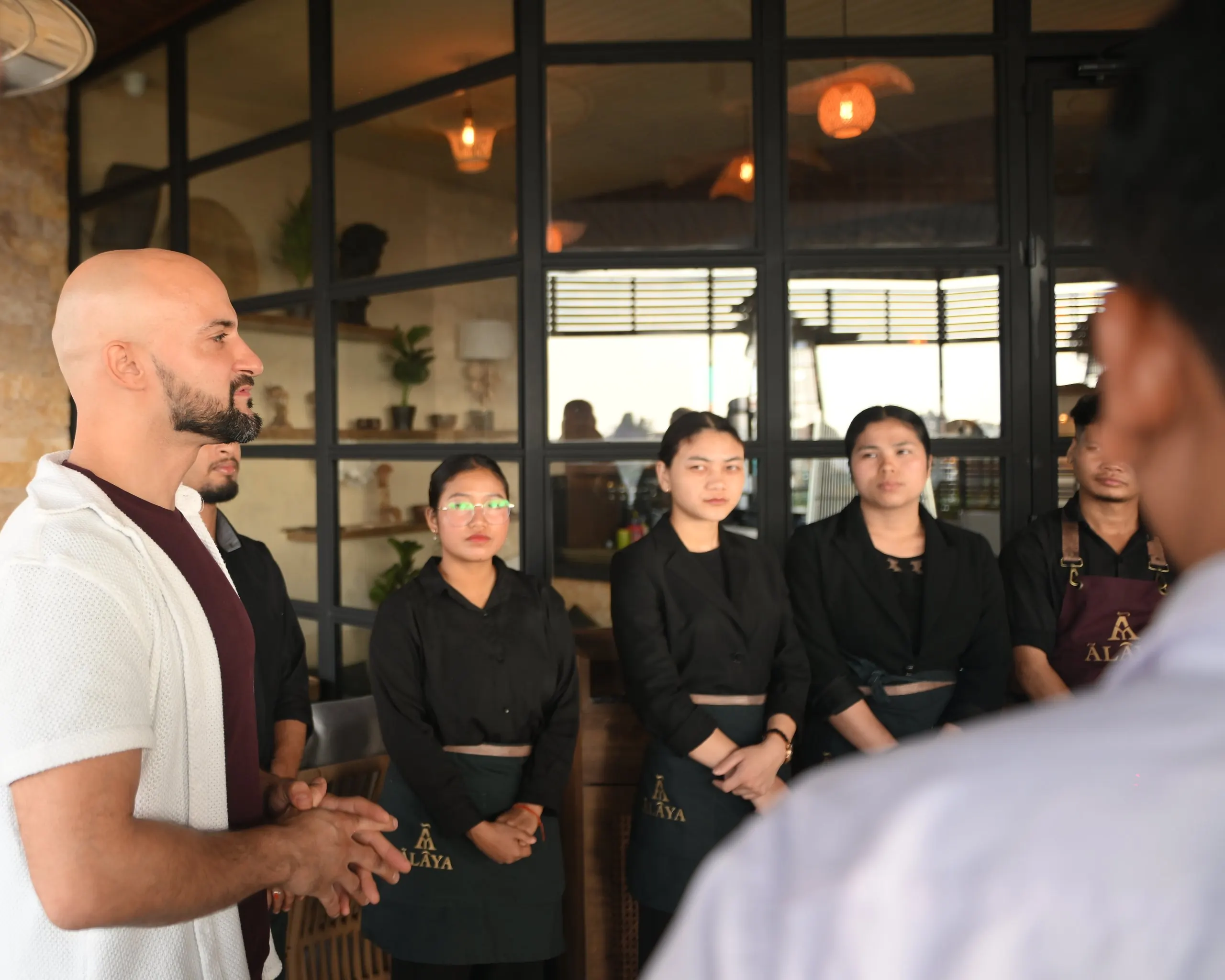 A briefing for the floor team on the Ālaya terrace by day: a man in a white overshirt speaks to staff in black uniforms and Ālaya aprons