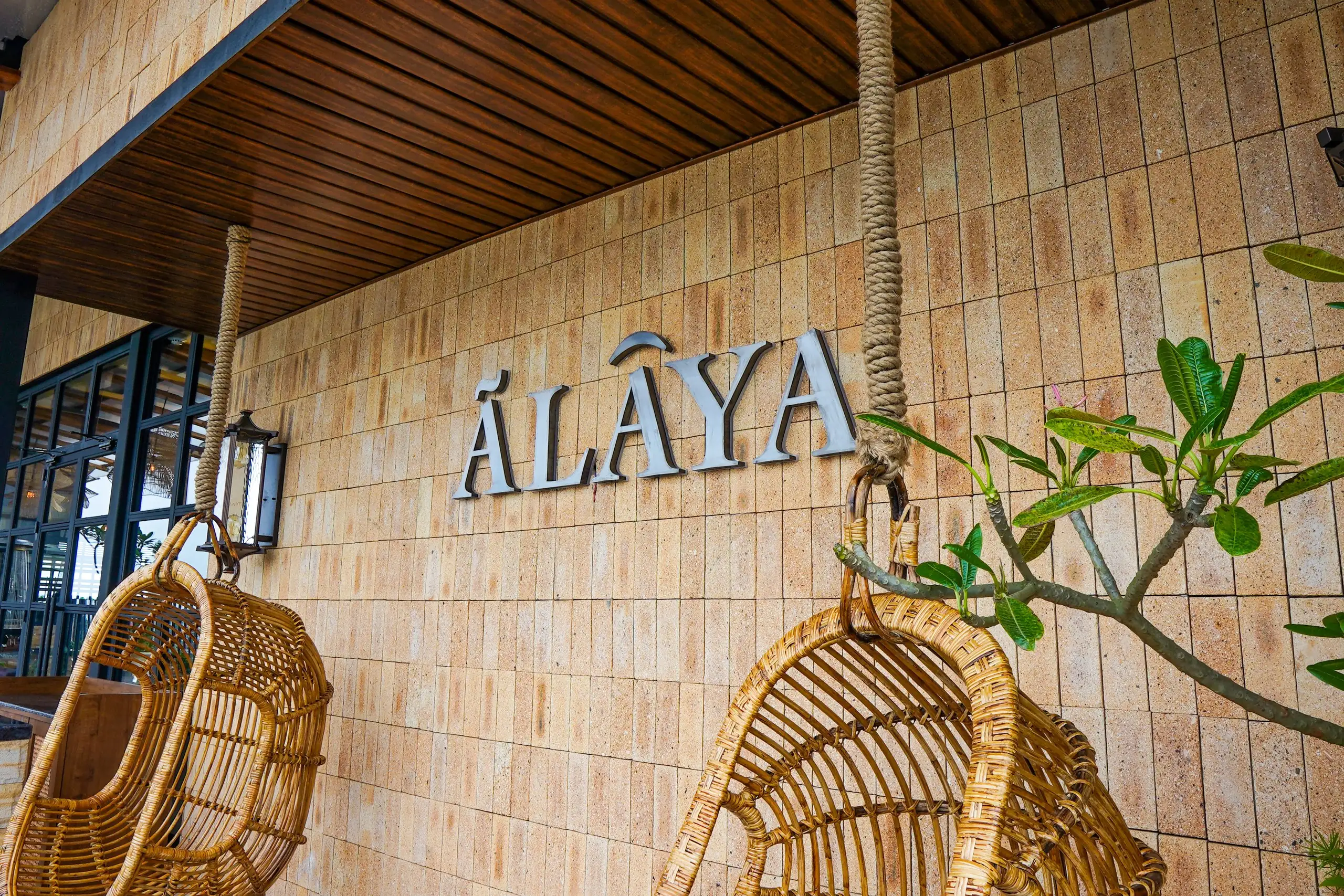 The Ālaya name in steel letters on the sandstone wall of its terrace by day, between two hanging rattan swing chairs and a frangipani