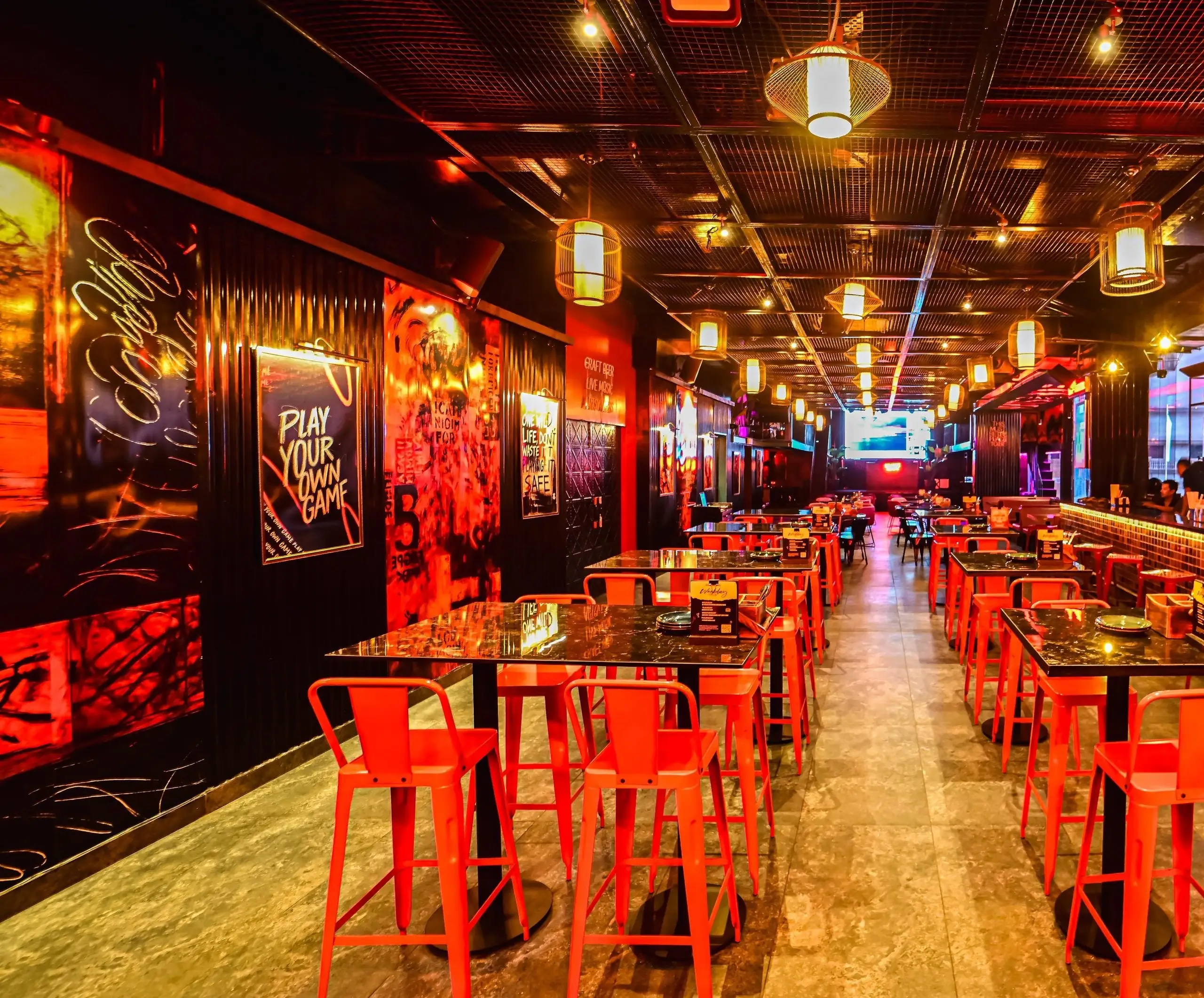 The repositioned hall at Nuts & Brew, Guwahati: red steel stools at black stone high tables under woven lanterns and a black mesh ceiling