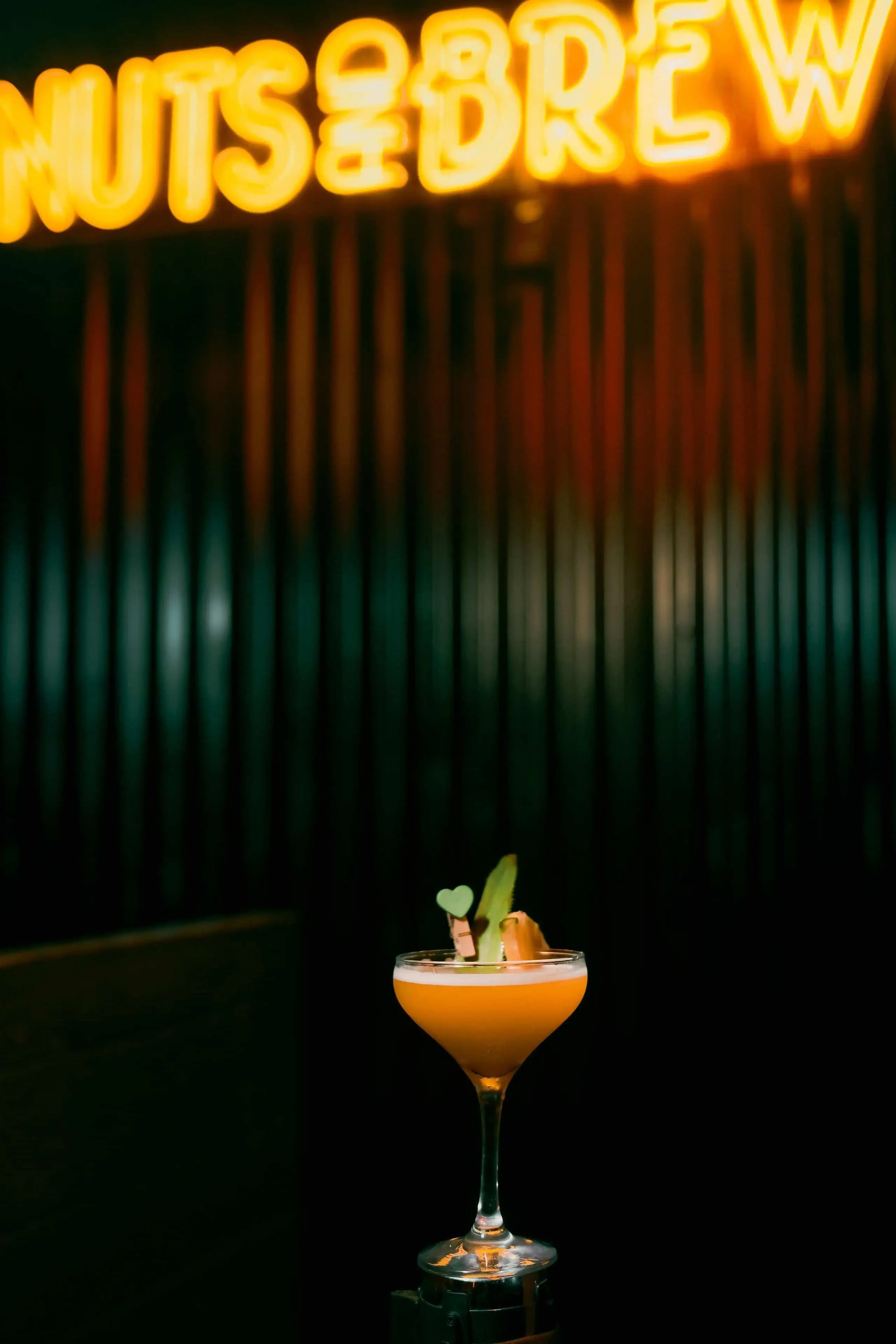 The Nuts & Brew neon sign glowing over a dark green corrugated wall, with a cocktail below