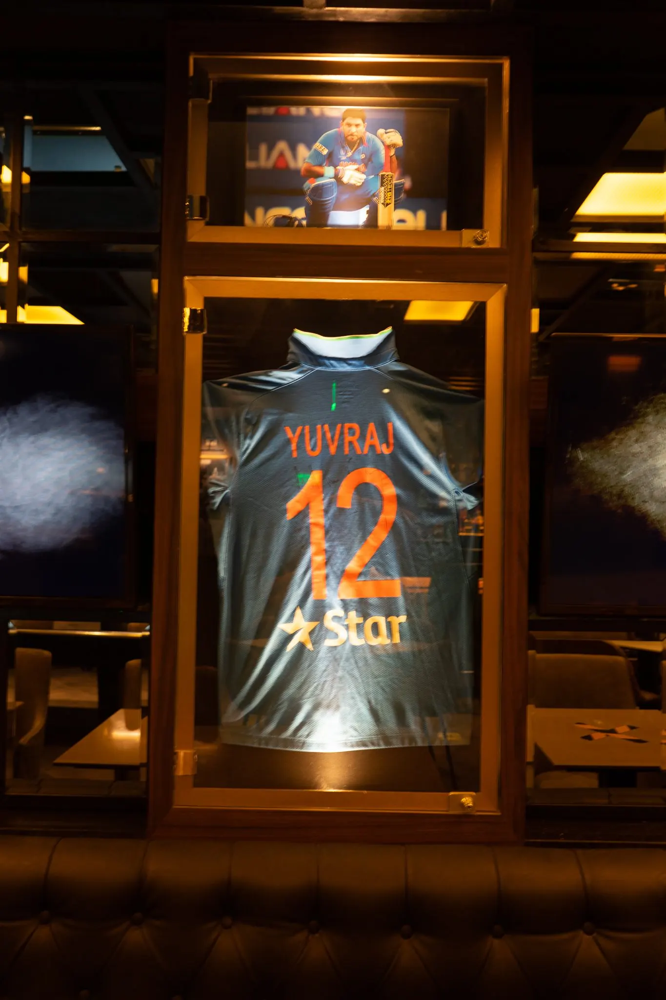 A framed cricket jersey in a timber display case above a leather banquette at Underdoggs Cyber Hub