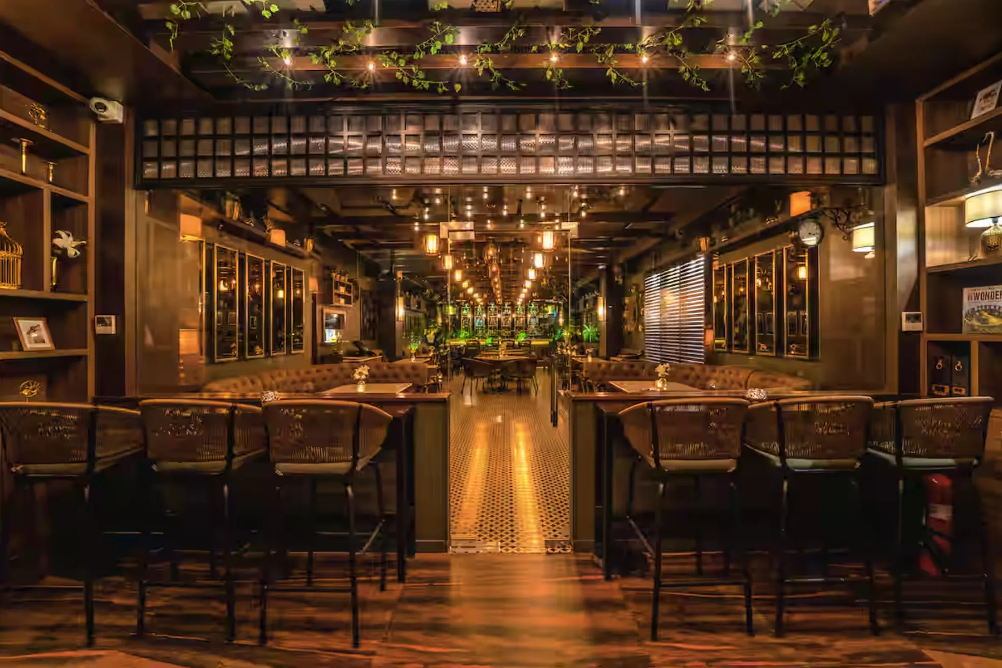 The central aisle at Uptown Escape, Guwahati: bar counters with woven high chairs either side, a patterned tile floor and a lattice ceiling hung with vines and lights