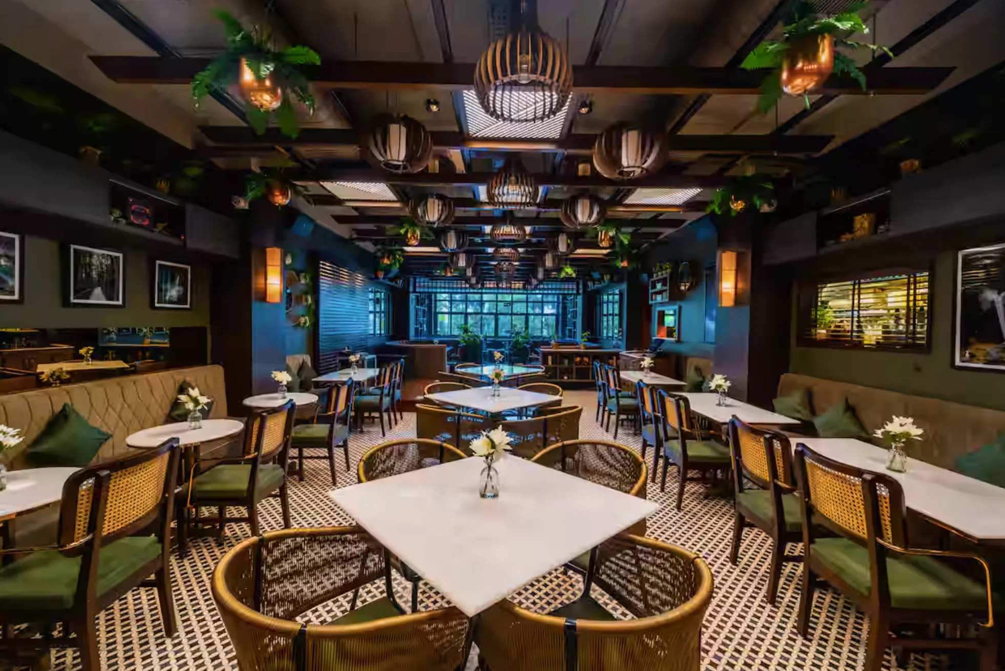 The dining room at Uptown Escape, Guwahati, in daylight: marble tables, rattan chairs, woven pendant lights and hanging plants below a window wall