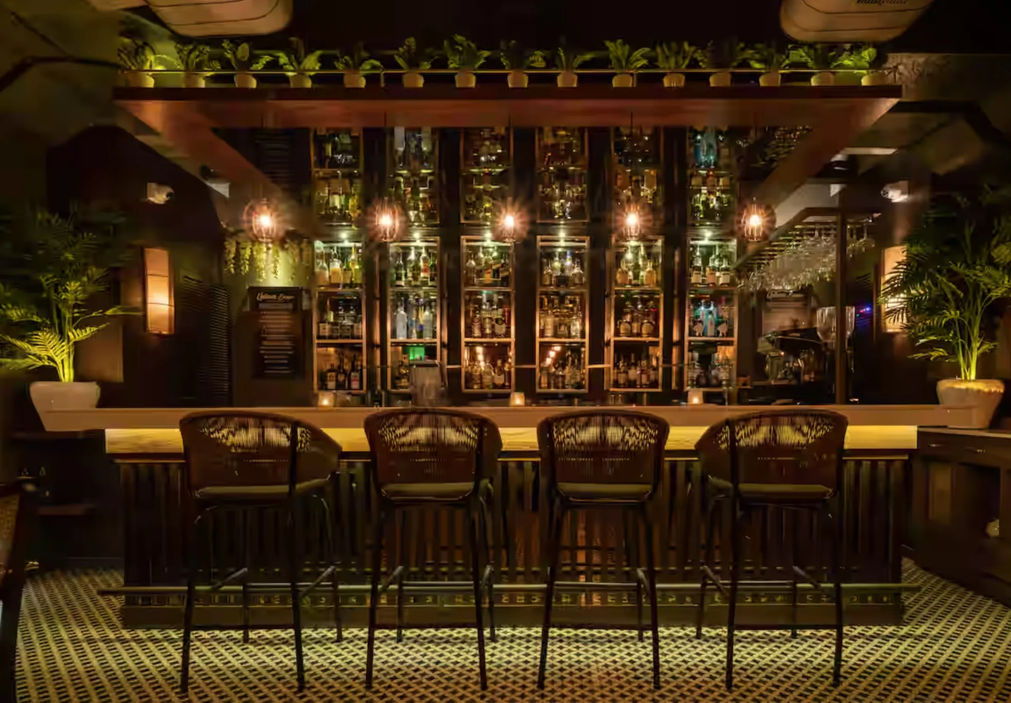 The same bar at Uptown Escape after dark: spirits on the lit back bar, woven high stools and planting along the gantry